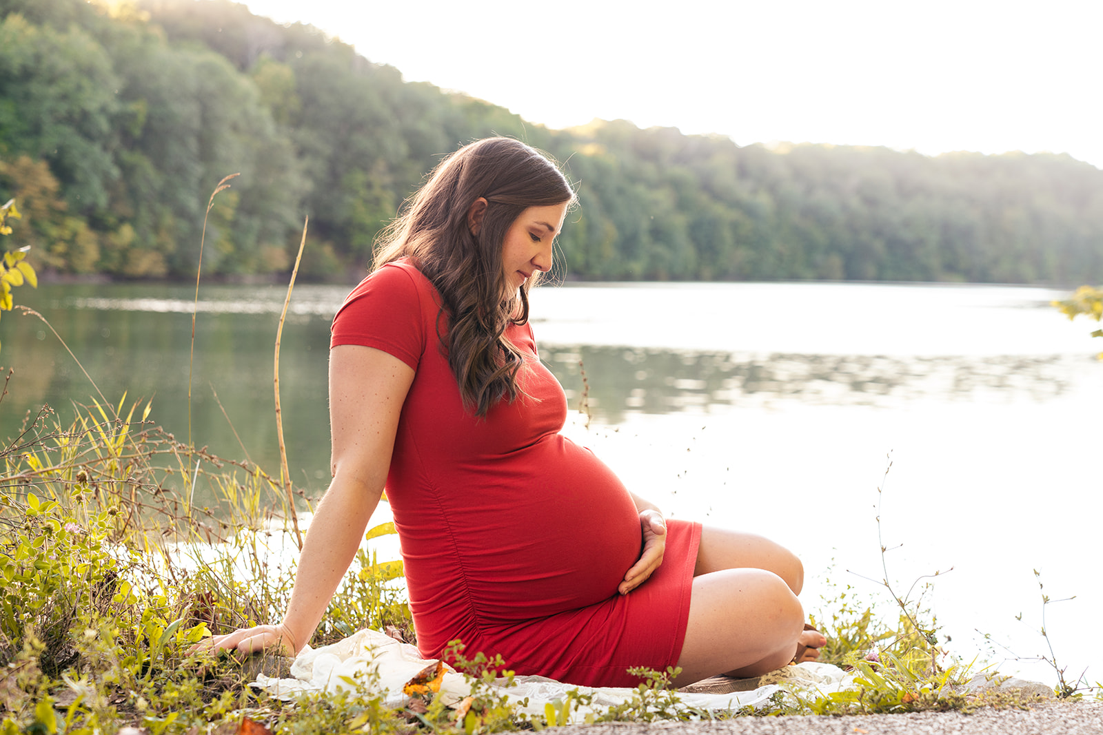 Mother looks down on pregnant belly with lake behind her